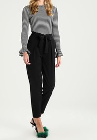 Svarta högmidjade byxor med knytband, kombinerade med en långärmad topp i svart och vitt randig design med trumpetärmar. Gröna klackar accentuerade med päls.