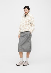 Νεαρή γυναίκα που φοράει μπεζ φλις με σχέδια, γκρι φούστα τύπου cargo, λευκές κάλτσες και ροζ ανοιχτά αθλητικά παπούτσια, στέκεται μπροστά σε λευκό φόντο.