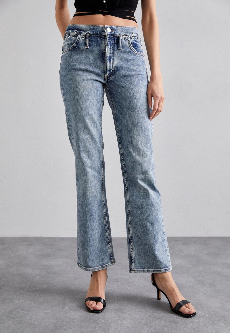 Jean en denim bleu clair avec jambes évasées, détails cousus et fermeture à boutons sur le devant. Associé à des sandales noires à talons hauts.