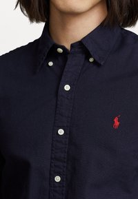 Marinblå knapp-up skjorta i bomull, med vita knappar och en röd broderad logotyp på vänster bröst. Slät textur och klassisk krage.