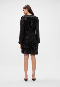 Vestido preto de lantejoulas com mangas longas, corpete texturizado e franzido, barra com volantes e silhueta ajustada, usado com sapatos de salto pretos.