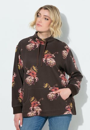 Kvinde med kort blondt hår iført en mørkebrun blomstret pullover sweatshirt med snore og blå jeans, stående foran en hvid baggrund.