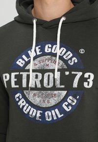 Sudadera con capucha verde oscuro con cordones blancos y un gran gráfico circular en azul, blanco y rojo, que dice "PETROL '73" y "CRUDE OIL CO."