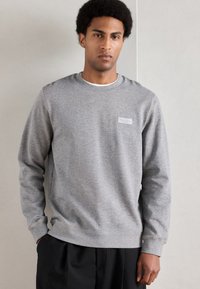 Sweatshirt cinzento com decote redondo, apresentando uma textura subtil e uma pequena etiqueta retangular no peito esquerdo; combinado com calças pretas.
