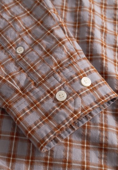 Poignet boutonné d'une chemise gris clair à motif à carreaux orange et blanc, montrant deux boutons blancs sur le poignet.