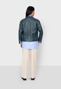 Tyrkysová kožená bunda s přiléhavým střihem, viditelným šitím a zadním ventilačním otvorem. Kombinována se světle modrou košilí a béžovými kalhotami.