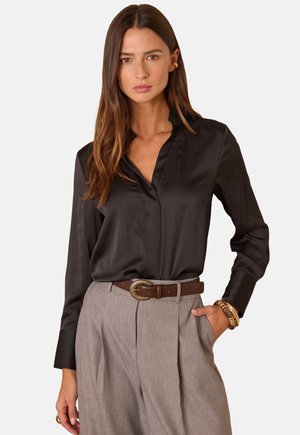Blusa in satin nero con colletto, maniche lunghe e texture liscia, abbinata a pantaloni grigi a vita alta e una cintura marrone con fibbia dorata.