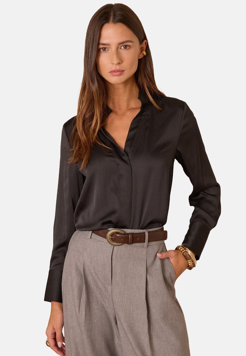 Blusa in satin nero con colletto, maniche lunghe e texture liscia, abbinata a pantaloni grigi a vita alta e una cintura marrone con fibbia dorata.