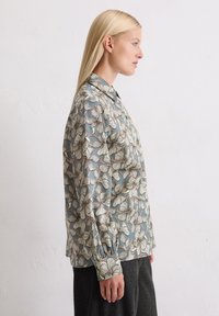 Floral patterned blouse in light blue with white and brown accents, featuring a collared neckline and long sleeves. Smooth texture.