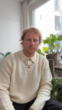 Krēmkrāsas, teksturēts adīts džemperis ar apkakli un pogu joslu, valkā sudraba ķēdi. Fonā redzami zaļi augi.