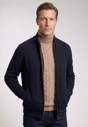 Mann trägt einen beigen, kabelgestrickten Rollkragenpullover und eine dunkelblaue Zip-up-Jacke mit den Händen in den Taschen vor einem einfarbigen Hintergrund.