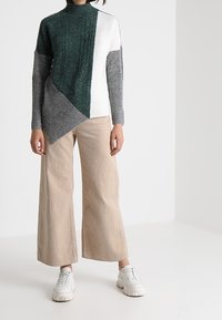 Pull en tricot vert foncé, blanc et gris avec un col montant et un motif texturé, associé à un pantalon beige à jambes larges et des baskets blanches.