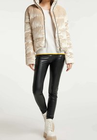Chaqueta beige acolchada con una textura brillante, capucha y cremallera frontal, combinada con pantalones negros ajustados y botas blancas de cordones gruesos.
