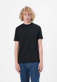Fekete pamut póló rövid ujjakkal és kerek nyakkal. Diszkrét logó hímezés található a mellkason. A modellen kék farmernadrág van.