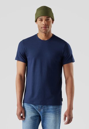 Mann trägt eine olivgrüne Strickmütze, ein marineblaues kurzärmeliges T-Shirt und hellblaue Jeans, steht vor einem hellgrauen Hintergrund.