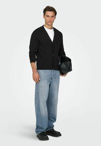 Cardigan nero lavorato a maglia, abbottonato, indossato sopra una t-shirt bianca. Jeans a gamba larga color azzurro chiaro e scarpe nere robuste, con una borsa nera in mano.