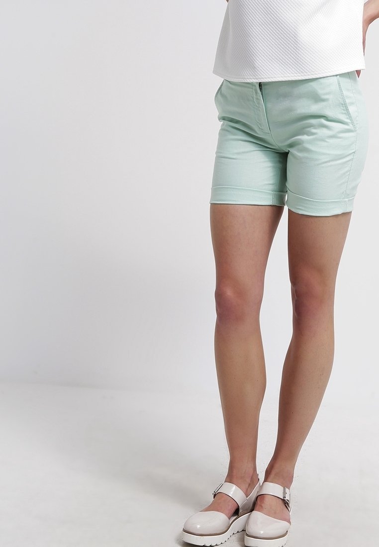 Shorts vert menthe clair en mélange de coton, dotés d'une fermeture éclair frontale et de revers roulés. Portés avec un haut blanc texturé et des chaussures claires.