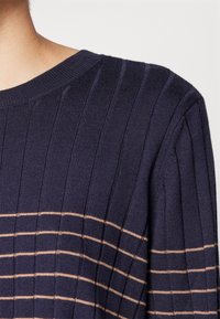 Granatowy sweter prążkowany z brzoskwiniowymi poziomymi paskami, z okrągłym dekoltem, noszony przez osobę o jasnej karnacji na białym tle.