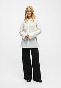 Chemise blanche à boutons et rayures verticales, à manches longues et avec un col, associée à un pantalon large noir et des chaussures noires.