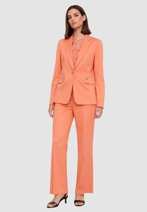 Donna in piedi che indossa un blazer sartoriale color pesca con pantaloni dritti abbinati e tacchi neri aperti, su sfondo neutro.