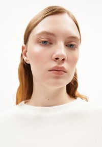 Nahaufnahme einer Person mit heller Haut und hellbraunen Haaren, die ein strukturiertes weißes Oberteil und kleine Steckerohrringe trägt. Neutrale Gesichtsausdruck.