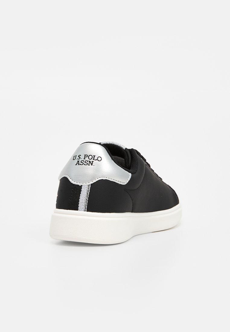 Zapatilla negra con un upper sintético liso, acento plateado en el talón y suela blanca. Presenta un logotipo sutil en la parte trasera y detalles a rayas.