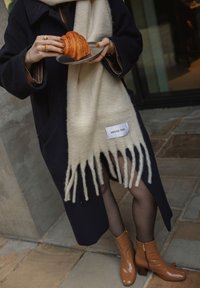 Donkerblauwe jas, beige sjaal met franjes met het label "ARCTIC FOX", houdend een croissant op een bord. Bruine enkellaarzen met een rits, doorzichtige panty's.