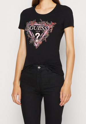 Frau trägt ein schwarzes Guess-T-Shirt mit einem floralen dreieckigen Logo-Design und schwarze High-Waist-Jeans, steht vor einem schlichten Hintergrund.
