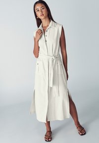 Donna che indossa un vestito camicia beige senza maniche con cintura, bottoni frontali, grandi tasche e spacchi laterali, abbinato a sandali neri.