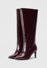 Une paire de bottes en cuir bordeaux foncé, montantes jusqu'au genou, avec des bouts pointus et des talons moyens fins sur fond blanc.