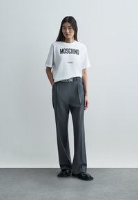 Biele bavlnené tričko s potlačou "MOSCHINO Milano", skombinované s vysokými šedými nohavicami na mieru a čiernymi topánkami.