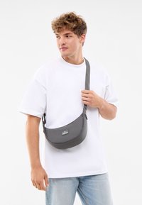 Graue Umhängetasche aus strukturiertem Stoff, halbkreisförmig mit einem langen Tragegurt und einem kleinen Markenlogo-Patch auf der Vorderseite.
