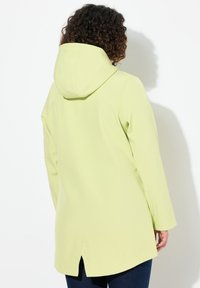Cappotto verde chiaro con cappuccio, maniche lunghe e fessure laterali all'orlo. Realizzato in un tessuto liscio e flessibile. Mostrata la vista posteriore.