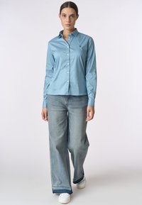 Camicia button-up azzurro chiaro realizzata in tessuto liscio, con colletto a punta e piccolo dettaglio ricamato. Abbinata a jeans a gamba larga sbiaditi e sneakers bianche.
