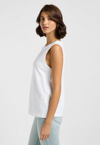 Top blanco sin mangas hecho de material suave, con un escote redondo y un corte relajado, combinado con jeans ajustados azul claro.