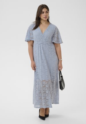 Femme portant une robe longue en dentelle bleu clair avec des manches évasées, tenant un sac à main noir et portant des talons noirs sur un fond uni.