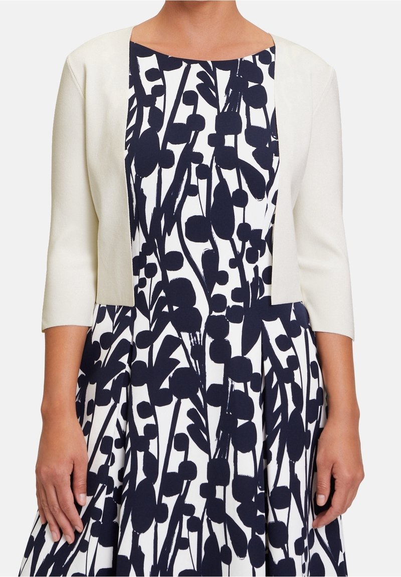 Femme portant une veste courte blanche par-dessus une robe blanche avec un motif floral abstrait noir audacieux, visible des épaules jusqu'au milieu des cuisses.