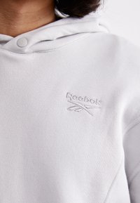 Világosszürke kapucnis pulóver, puha anyagból készült, elöl kenguru zsebből és hímzett Reebok logóból áll. Állítható zsinóros kapucnival rendelkezik.