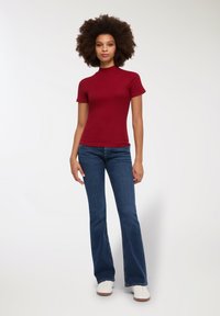 Maglione rosso a maniche corte con collo alto abbinato a jeans a campana blu. Il top ha una texture liscia; i jeans presentano una finitura denim classica. Sneakers bianche completano il look.