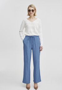 Top in pizzo bianco a maniche lunghe con scollatura a V, abbinato a pantaloni larghi blu chiaro e scarpe con tacco marroni. L'outfit include occhiali da sole.