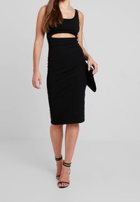 Musta hihaton midi-mekko, jossa on leikkauksellinen yksityiskohta vyötäröllä, istuva siluetti ja teksturoitu kangas; yhdistetty mustiin remmissandaaleihin.