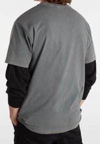 Camiseta de algodón gris con un ajuste relajado, cuello redondo y mangas cortas. La vista posterior muestra una textura suave y costuras sutiles, superpuesta sobre mangas largas negras.