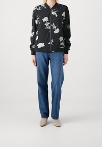 Kvinna klädd i svart blommig zip-up jacka, blå raka jeans och beige ankelskor mot en enfärgad bakgrund.
