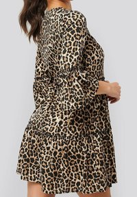 Kleid mit Leopardenmuster, das ein geschichtetes Design, Puffärmel und elastische Bündchen aufweist. Hergestellt aus einem glatten Stoff in Beige- und Schwarzton.