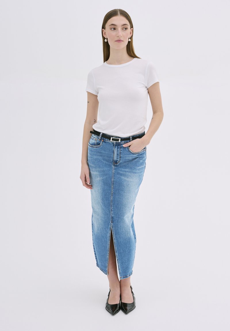 Młoda kobieta w białej koszulce z krótkim rękawem, w niebieskiej dżinsowej spódnicy midi z wysokim stanem i rozcięciem z przodu, z czarnym paskiem i czarnymi spiczastymi butami, stojąca na białym tle.