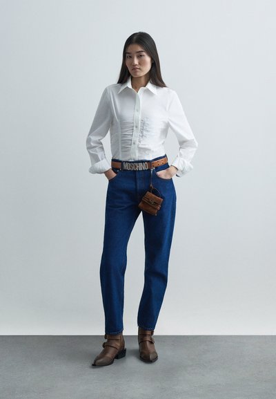 Camicia bianca a maniche lunghe con arricciatura frontale, pantaloni in denim blu, cintura marrone con fibbia "MOSCHINO", piccola borsa marrone, scarpe marroni.