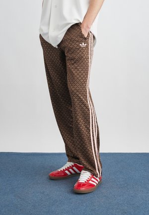 Άτομο που φορά καφέ παντελόνι φόρμας Adidas με ρόζ ρίγες και κόκκινα παπούτσια Adidas, στέκεται σε μπλε χαλί.