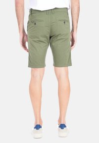 Pantaloni corti verdi in cotone con un taglio dritto, tasche anteriori e posteriori e una cintura. Un modello indossa scarpe da ginnastica casual.