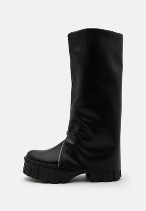 Botte haute en cuir noir avec semelle épaisse et rainurée et fermeture éclair verticale sur le côté avant, sur un fond uni.