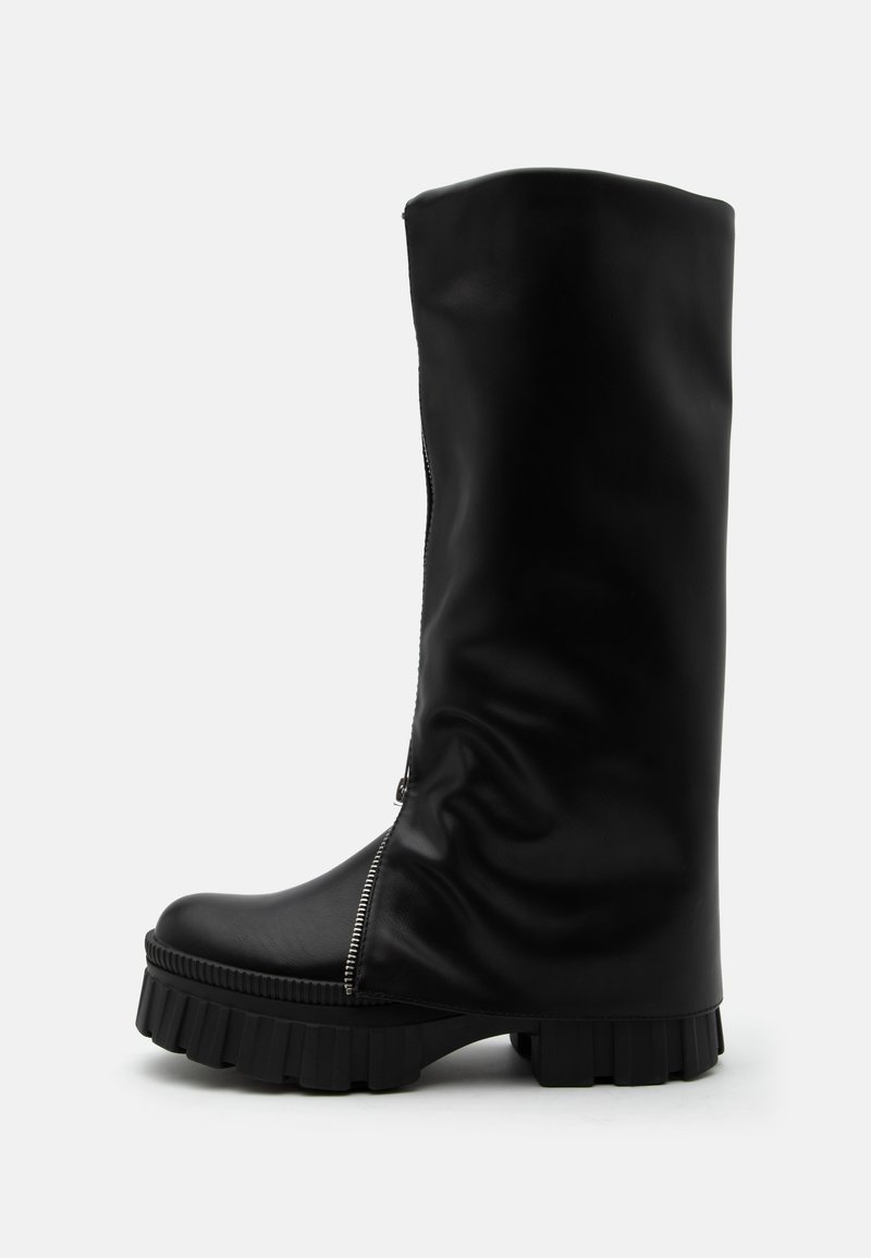Botte haute en cuir noir avec semelle épaisse et rainurée et fermeture éclair verticale sur le côté avant, sur un fond uni.